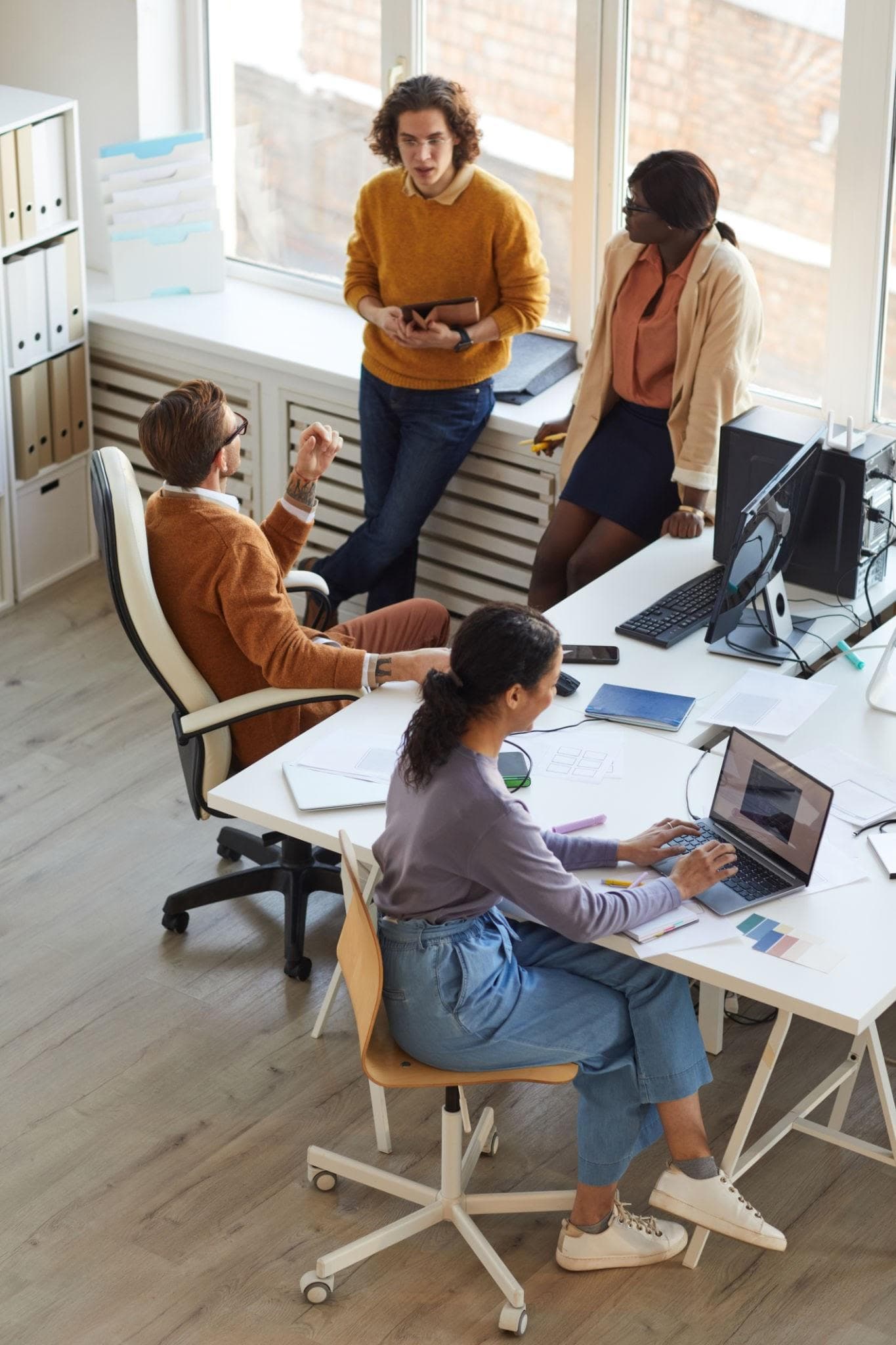 Equipe conversando em um escritório: duas pessoas em pé junto à janela e duas sentadas às mesas de trabalho